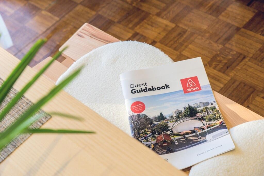 Guide de bienvenue Airbnb posé sur une table en bois clair, avec une plante verte en avant-plan et un parquet en arrière-plan. La couverture du guide affiche une image de paysage urbain avec le logo Airbnb.