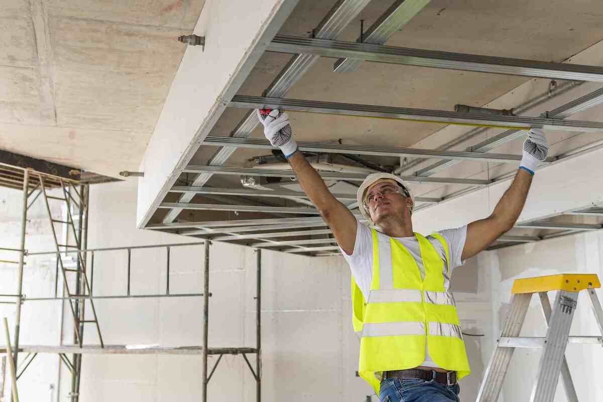 Un artisan travaille sur l'installation d'une structure métallique au plafond d'un bâtiment en construction. Équipé d'un casque et d'un gilet de sécurité, il mesure avec précision pour garantir un résultat fiable et solide, renforçant sa réputation professionnelle dans le secteur des travaux.